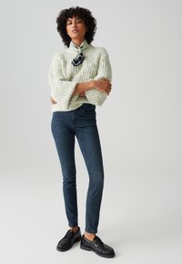Frau mit lockigem Haar steht mit verschränkten Armen, trägt einen hellgrünen grob gestrickten Pullover, dunkle enge Jeans, schwarze Schuhe und ein Halstuch.