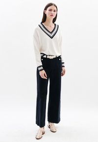 Maglione crema con scollo a V a righe blu navy e polsini, abbinato a pantaloni a gamba larga blu navy scuri a vita alta e scarpe strappy beige chiaro.