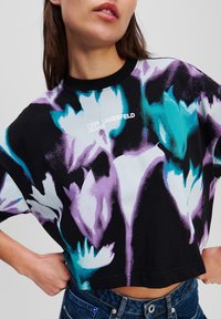 Juoda trumpa palaidinė su mėlynojo ir levandų atspalvio gėlių raštu, o priekyje yra tekstas "KARL LAGERFELD JEANS" baltos spalvos.