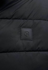 Schwarze Pufferjacke aus strukturiertem Stoff, mit gesteppten Abschnitten und einem auffällig platzierten schwarzen Gummilogo-Patch.