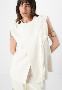 Cream-colored sleeveless top with a diagonal fringe detail along the side, featuring a round metallic accent at the shoulder. Smooth texture.