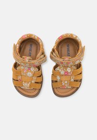 Geel suède sandalen hebben bloemenpatroonaccenten, meerdere verstelbare banden en een bruine gestructureerde zool. Merknamen zichtbaar aan de binnenkant.