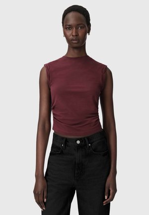 Frau steht vor schlichtem Hintergrund, trägt ein ärmelloses bordeauxfarbenes Top und hoch taillierte schwarze Jeans und schaut mit neutralem Gesichtsausdruck nach vorne.