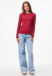 Jongeman staat, gekleed in een rode langwerpige shirt, lichtblauwe wijde jeans en beige sneakers, tegen een effen witte achtergrond.