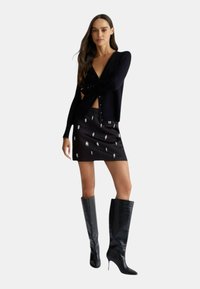 Cardigan nero a coste con bottoni, abbinato a una mini gonna nera con dettagli argento e stivali neri lucidi fino al ginocchio.