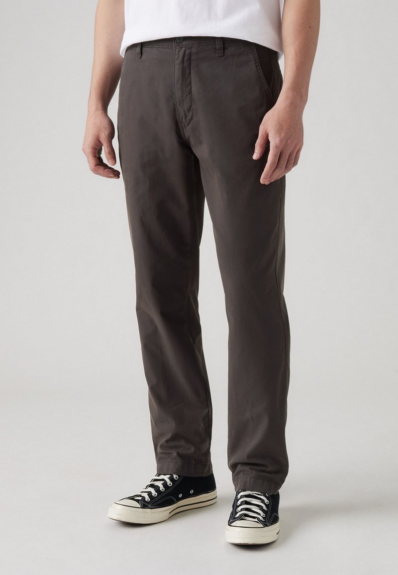 Pantalones de color marrón oscuro, hechos de algodón, con un diseño de pierna recta, bolsillos frontales y una textura suave. Combinados con zapatillas negras.