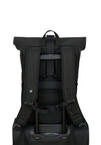 Un sac à dos noir avec des bretelles rembourrées attaché à la poignée et au châssis d'une valise à roulettes, vu de l'arrière sur un fond blanc.