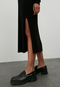 Schwarzes geripptes Midi-Kleid mit einem seitlichen Schlitz, kombiniert mit klobigen schwarzen Slippern aus glattem Leder und dicker Sohle.
