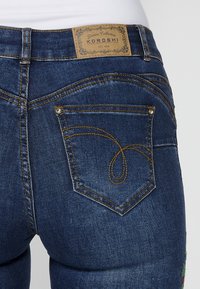 Dark blue denim jeans with a textured finish, featuring gold stitching and a heart-shaped design on the back pocket. Leather brand tag visible.