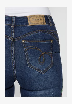 Jeans in denim blu scuro con una finitura testurizzata, caratterizzati da cuciture dorate e un design a forma di cuore sulla tasca posteriore. Etichetta del marchio in pelle visibile.