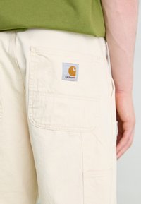 Pantalons cargo couleur crème avec de multiples poches, ornés d'un patch logo Carhartt en orange et gris à l'arrière. Tissu en coton, design décontracté.