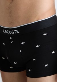 Črne bombažne bokserice z večkratnim vzorcem belega krokodiljega logotipa in črnim pasom z sivo obrobo, označene z "LACOSTE."