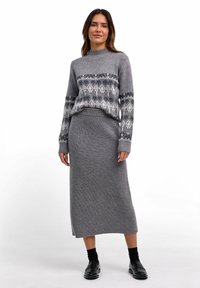 Frau, die einen grauen, gemusterten Strickpullover und einen passenden gerippten Strick-Midirock trägt, mit schwarzen Socken und schwarzen Loafers, stehend vor einem weißen Hintergrund.