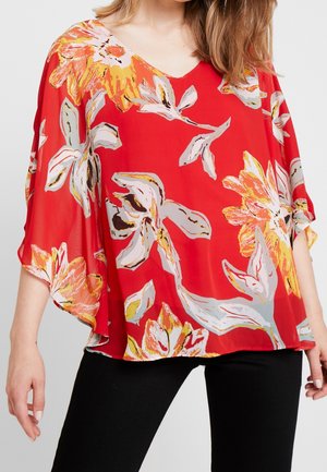 Kvinde iført løs rød bluse med store gule, hvide og grå blomsterprint, kombineret med sorte tætsiddende bukser.