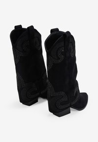 Botas de vaquero negras de ante con punteras afiladas. Presentan tachuelas decorativas en patrones ondulados a los lados. Tacones cuadrados gruesos.