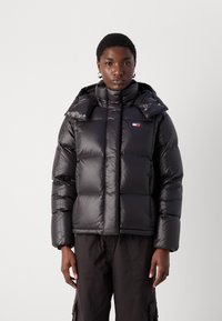 Juoda pūkinė striukė su blizgiu paviršiumi, turinti horizontalius susiuvus, aukštą apykaklę ir mažą logotipą ant krūtinės.