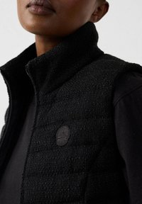 Gilet nero con texture e colletto alto, dotato di chiusura a zip e di un piccolo logo circolare sul lato sinistro. Indossato sopra un top scuro.