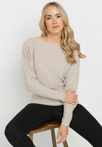 Beige ribbed knit sweater with a wide neckline and long sleeves, paired with black fitted pants. Wood stool adds a natural accent.
