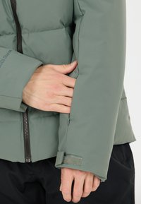 Giacca imbottita in verde tenue, con zip frontale, polsini regolabili con chiusura a Velcro e una finitura liscia e opaca.
