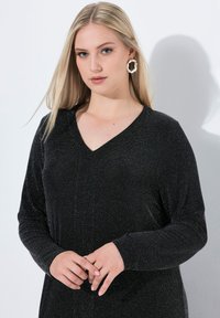 Schwarzes V-Ausschnittkleid mit langen Ärmeln; verfügt über eine glitzernde Textur und einen leichten Glanz. Die Modelf trägt große kreisförmige Ohrringe und hat die Hände vor sich gefaltet.
