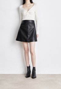Top bianco a maniche lunghe con un dettaglio frontale annodato, abbinato a una gonna a tubino nera in pelle e stivali neri alla caviglia. Texture lisce in tutto.