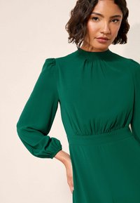 Zelené dlouhé šaty s dlouhým rukávem vyrobené z hladkého materiálu. Mají vysoký límec, nařasené rukávy a přiléhavý pas s jemným zdobením sklady.