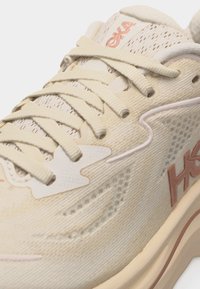 Beige og rosa Hoka løpesko med mesh-overdel, tykk såle og detaljert snøring på hvit bakgrunn.
