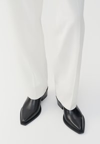 Bottines noires à bout pointu en cuir lisse, avec des talons bas en bloc, associées à un pantalon large blanc. Design simple et moderne.