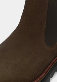 Botină ankle din suede maro cu o textură netedă, panouri laterale elastice și o talpă neagră robustă cu un detaliu de cusătură maro contrastant.