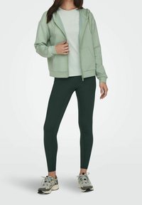 Světle zelená mikina s zipem přes bílý tričko, kombinovaná s tmavě zelenými legínami a šedými teniskami s zelenými akcenty. Hladk�á látka, volný střih.