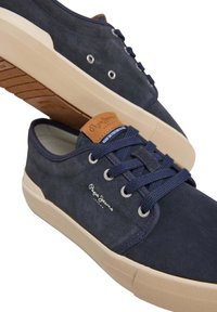 Sneakers scamosciati blu navy con suole in gomma beige, caratteristiche di accenti in pelle color cuoio, occhielli rotondi e lacci piatti blu navy. Logo su un lato.