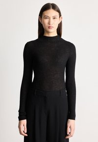 Jauna sieviete ar taisniem tumšiem matiem valkā melnu ribotu augsta kakla garu piedurkņu kreklu un vaļīgas melnas bikses, stāvot pret vienkāršu fonu.