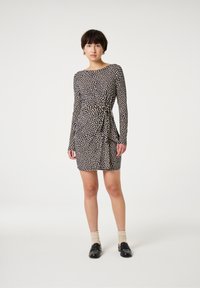 Schwarzes Kleid mit langen Ärmeln, gepunktetes Muster in Creme, figurbetonte Silhouette, Taillengürtel-Detail, knielange Länge, kombiniert mit schwarzen Slippern.