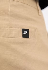 Béžové Nike kalhoty s malou černou nášivkou, jemnou mřížkovou texturou a diskrétními švy v oblasti kapes.
