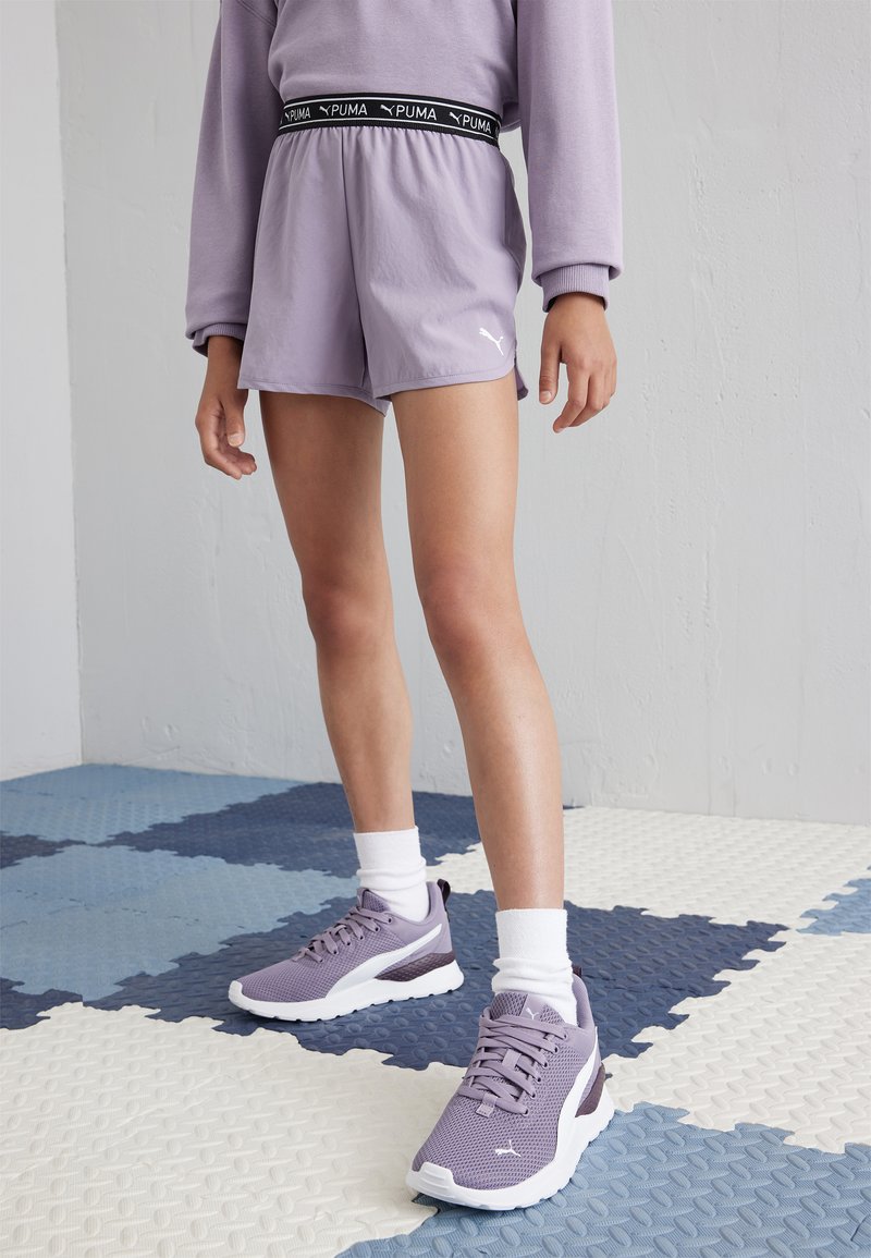 Pantalones cortos deportivos morados con una cinturilla negra de marca, combinados con zapatillas moradas que presentan detalles blancos y tela de malla, colocados sobre una esterilla texturizada.