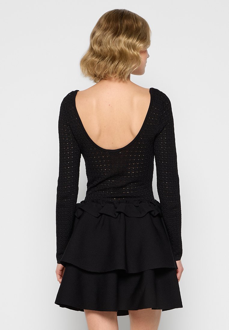 Schwarzes Kleid mit einem strukturierten, langärmeligen Oberteil mit tiefem Rückenausschnitt und einem geschichteten Volantrock. Der Stoff wirkt weich und strukturiert.