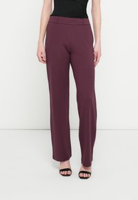 Maroonfarbene, weit geschnittene Hosen mit glatter Textur und tailliertem Bund. Das Modell trägt schwarze, offene Sandalen mit Absatz.