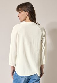 Chemise légère en crème texturée avec des manches trois-quarts, un ourlet arrondi et une coupe décontractée. Associée à un pantalon en jean, mettant en avant un style décontracté.