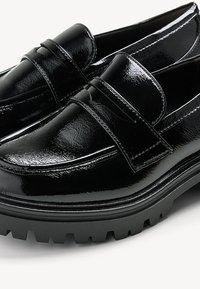 Mocassins en cuir verni noir avec un bout arrondi, dotés d'une bride traversant le cou-de-pied et d'une semelle extérieure noire épaisse et texturée.