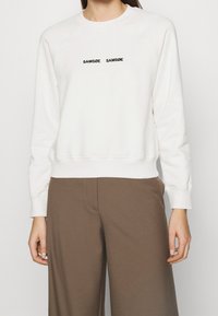 Sudadera color crema con cuello redondo, mangas raglán y dobladillo acanalado. Presenta texto negro: "SAMØE SAMØE" en el pecho.