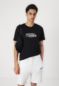 Svart t-skjorte med hvit "JACK & JONES" logo, kombinert med lysegrå shorts. Modellen bærer en svart crossbody-veske. Enkelt design, myk stoff.