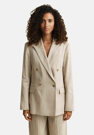 Vrouw met krullend haar, gekleed in een beige dubbelgeknoopte blazer met krijtstrepen en bijpassende broek, tegen een effen lichte achtergrond.