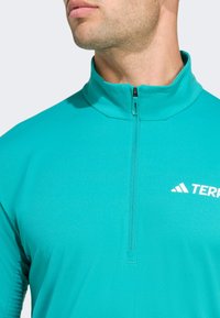 Tyrkysový sportovní top na zip s vysokým límcem, vyrobený z hladké, elastické tkaniny a s bílým logem na hrudi.