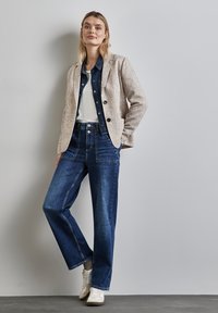 Ein hellbeiger, strukturierten Blazer über einem Jeanshemd, kombiniert mit hochgeschnittenen dunkelblauen Jeans und weißen Sneakers, schafft einen lässigen Schichtenlook.