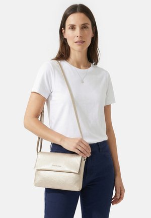 Frau mit langen braunen Haaren, die ein weißes T-Shirt und dunkle Hosen trägt, eine beige Umhängetasche und eine silberne Halskette.