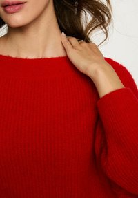 Maglione rosso a maglia con scollo ampio e maniche lunghe. Il materiale appare morbido e strutturato, con un motivo a coste.