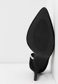 Chaussure à talon haut en daim noir à bout pointu avec semelle texturée, vue de dessous, appuyée verticalement contre un mur blanc.