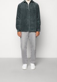 Persona che indossa una camicia di cotone a coste verde scuro sopra un dolcevita beige, jeans grigio chiaro e sneaker bianche, in piedi contro uno sfondo neutro.
