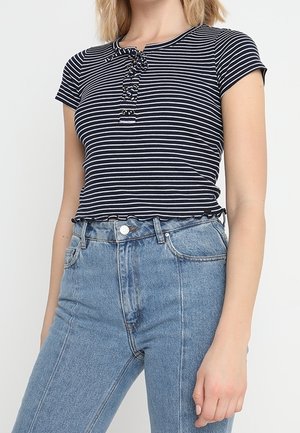 Kvinde iført en sort- og hvidstribet kortærmet top med knapper foran og højtaljede, lyseblå denimjeans.