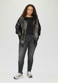 Zwart faux lederen shirtjack met twee voorkepers, gedragen over een zwart gebreid sweatervest en gecombineerd met donkere jeans en zilveren sneakers.